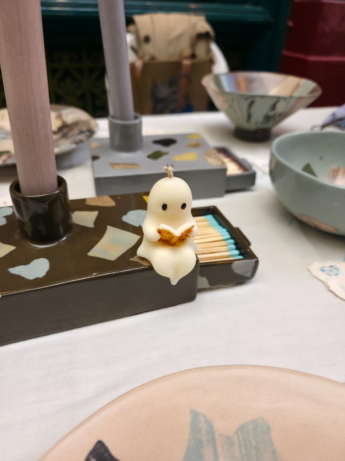 Little Ghost Reading, a small cream candle holding an open gold-edged book, perched on the corner of a dark glazed ceramic matchbox filled with teal-tipped matches.