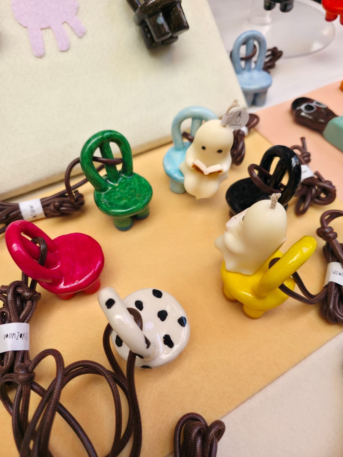 The same ghost candle sitting in a tiny pale-blue ceramic chair, surrounded by more chair-shaped ceramic pendants on brown cords in green, magenta, black, yellow and spotted white.